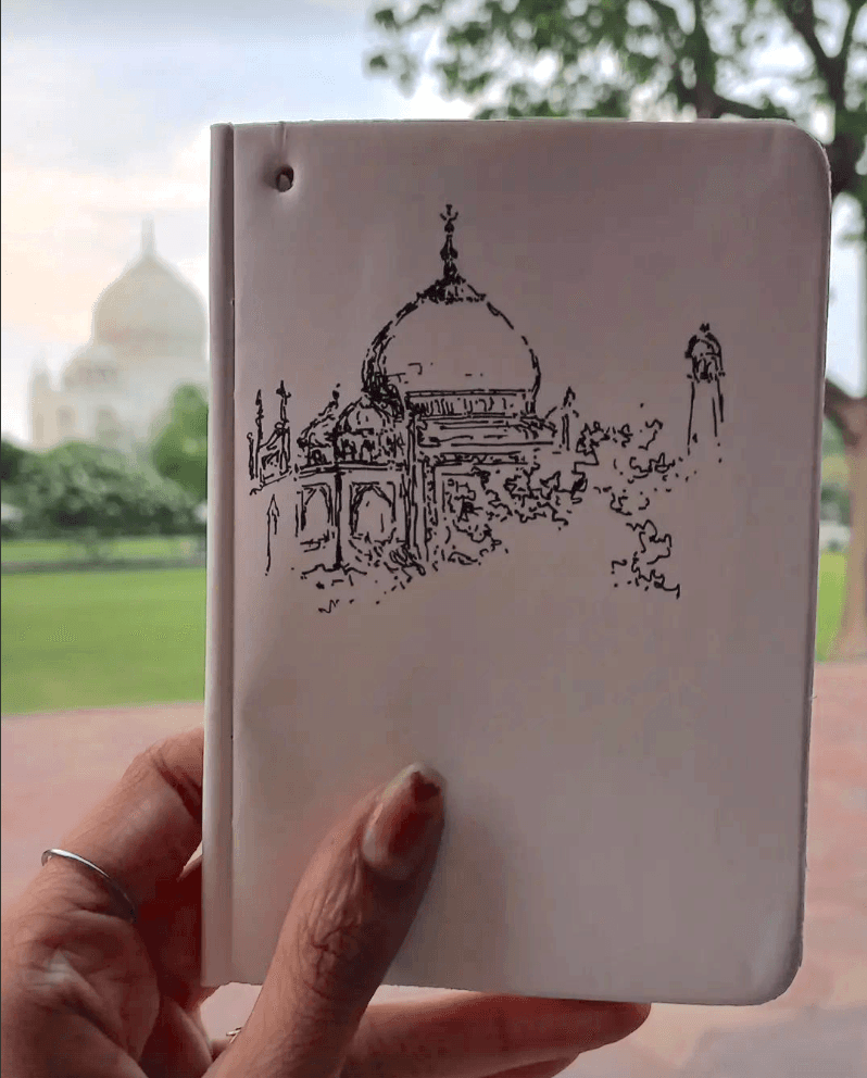 Hand holding a sketchbook with an ink drawing of the Taj Mahal, the real monument blurred in the background