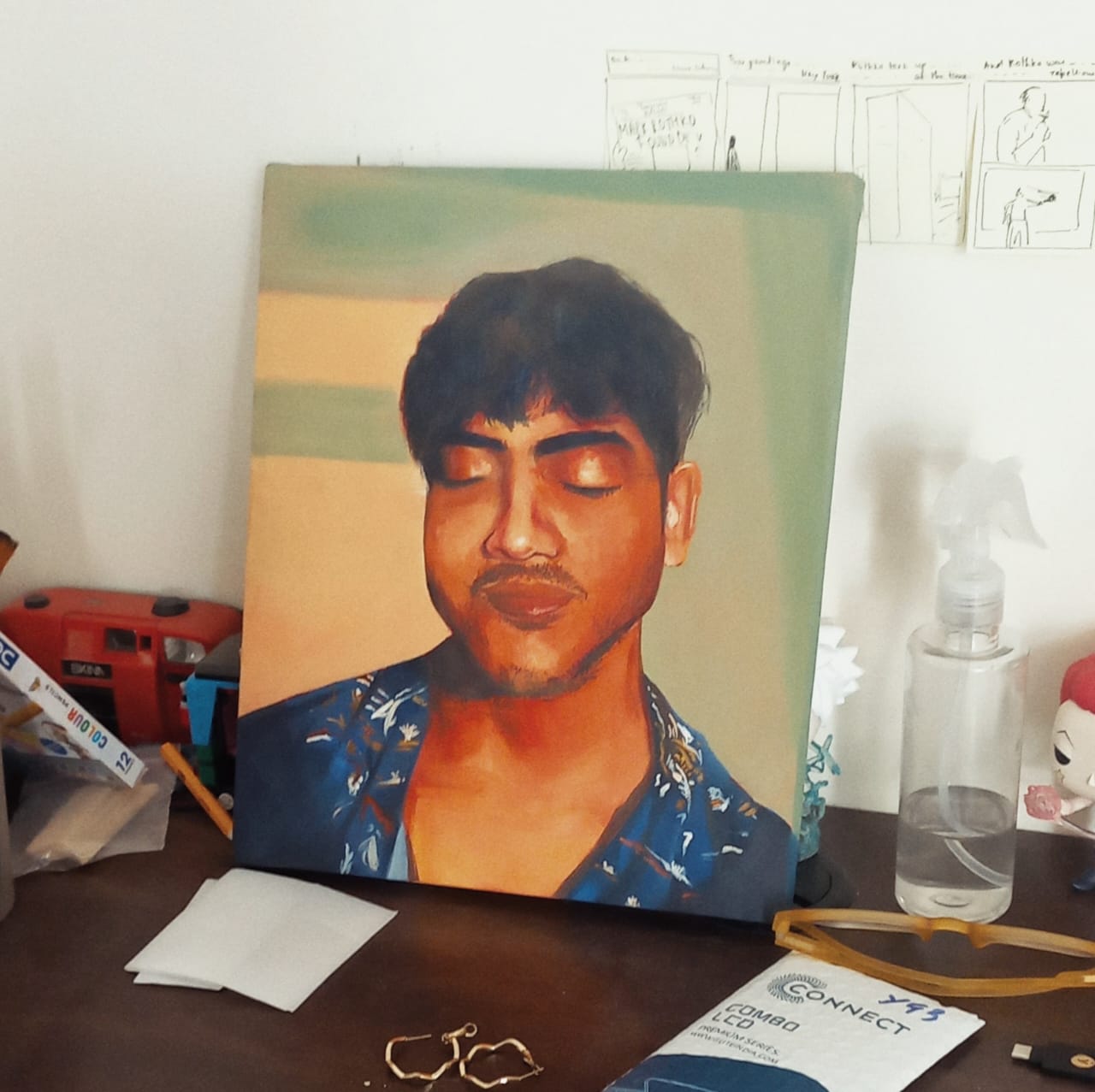 Painted portrait of a young man with closed eyes in a blue floral shirt, the canvas propped up on a desk