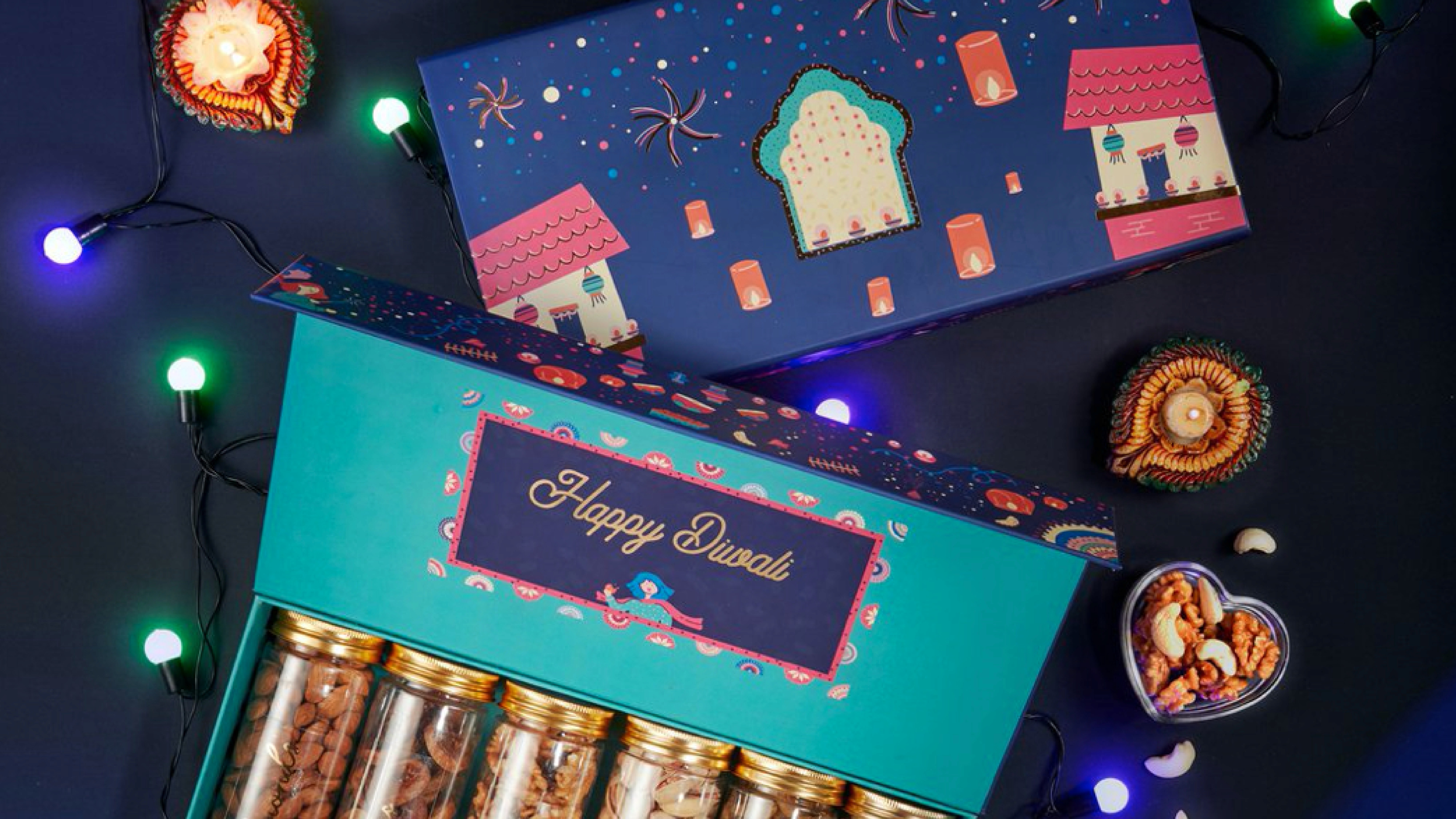 Native Picks Diwali dry fruit gift boxes in teal and navy, illustrated with village houses, lanterns and fireworks, among diyas and fairy lights