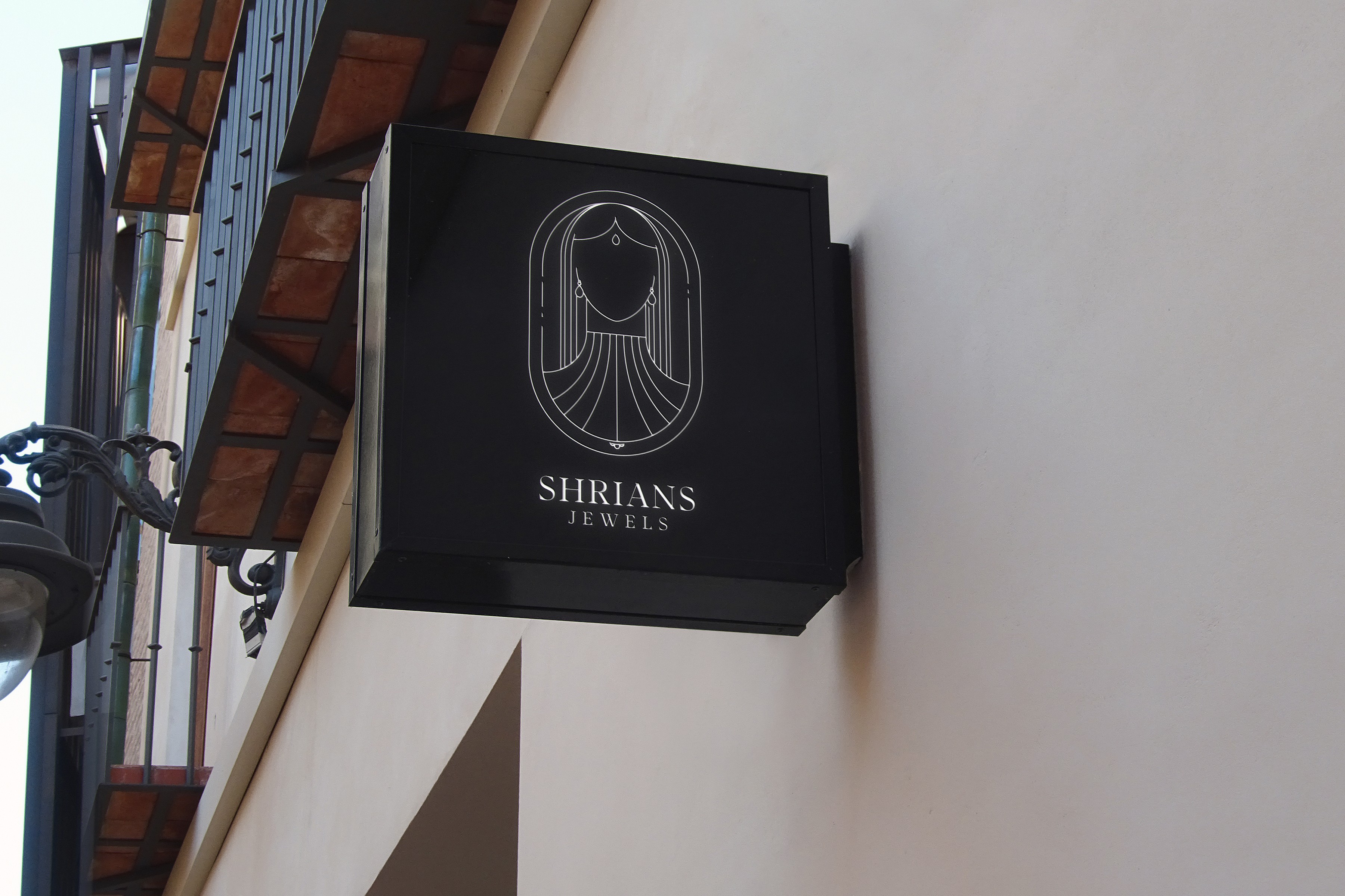Shrians Jewels shop sign mockup: a copper line-art emblem of a bride in an arch on a black sign
